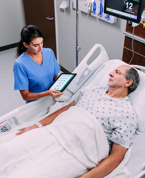 Female nurse consults tablet device at hospital bedside of older male patient